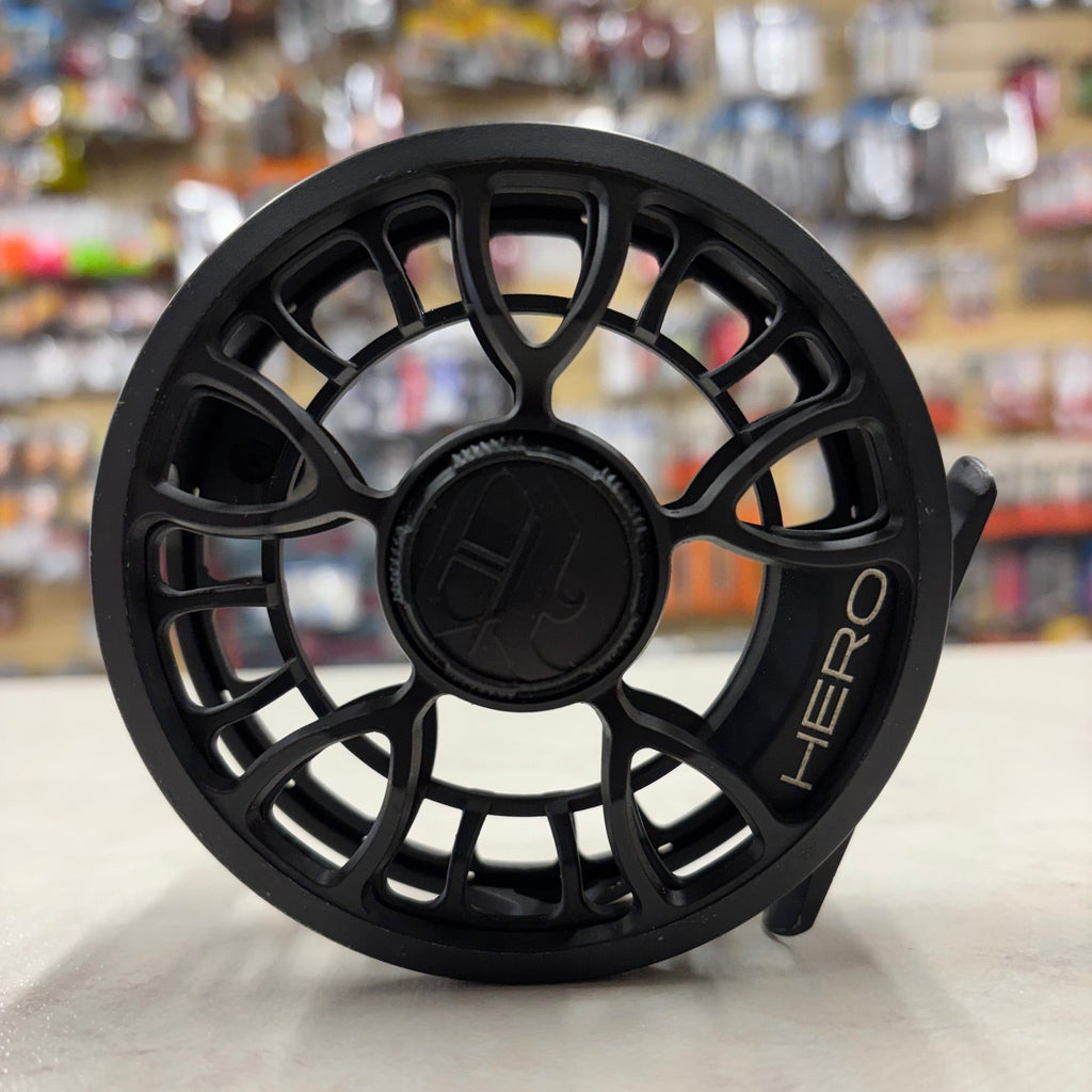 A black fly fishing reel with a textured, open-frame design and the brand name 'HERO' printed on the side, set against a blurred background of a retail store.