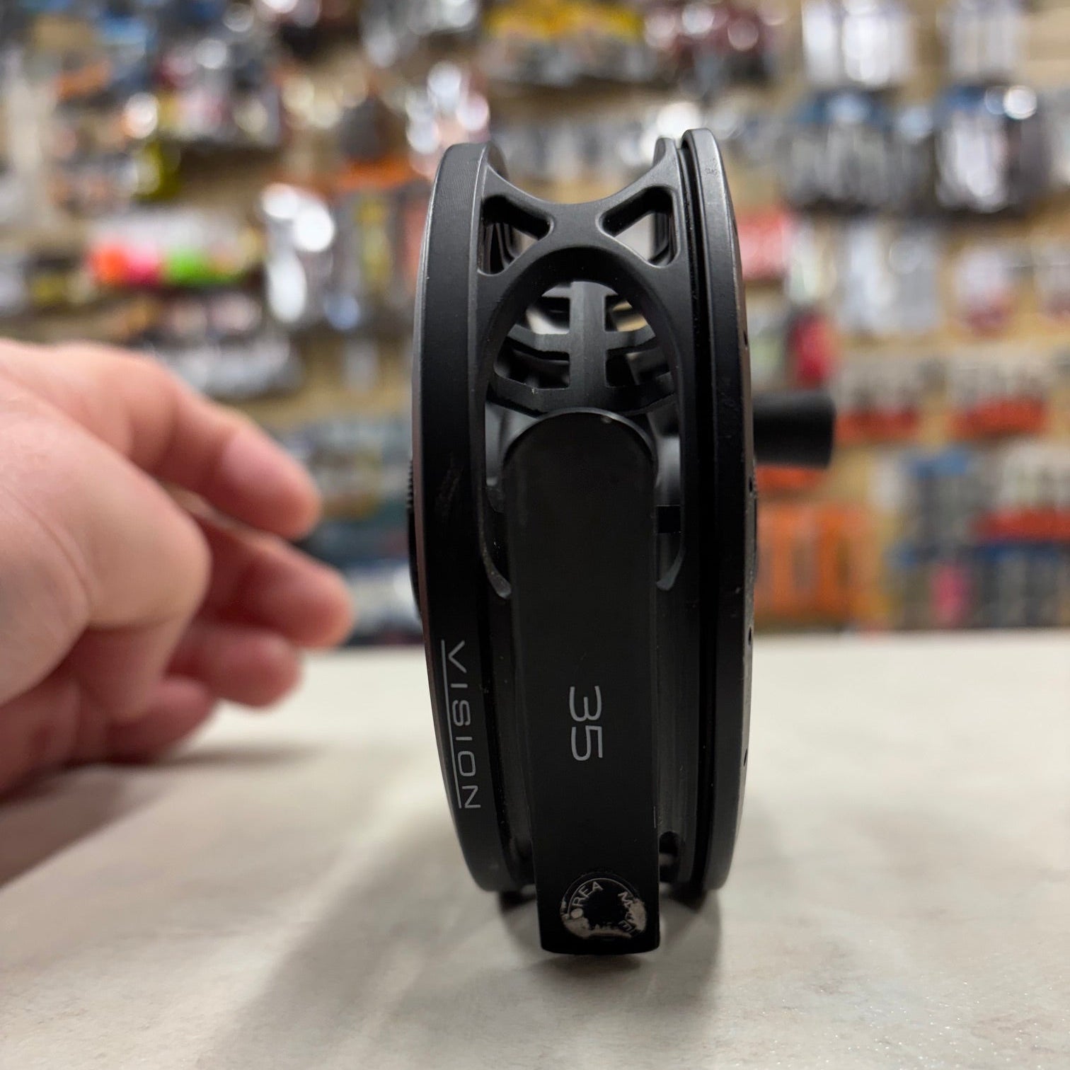 A close-up of a black fly fishing reel, model Vision 35, resting on a light gray surface with a hand pointing towards it. The background is a blurry store interior.