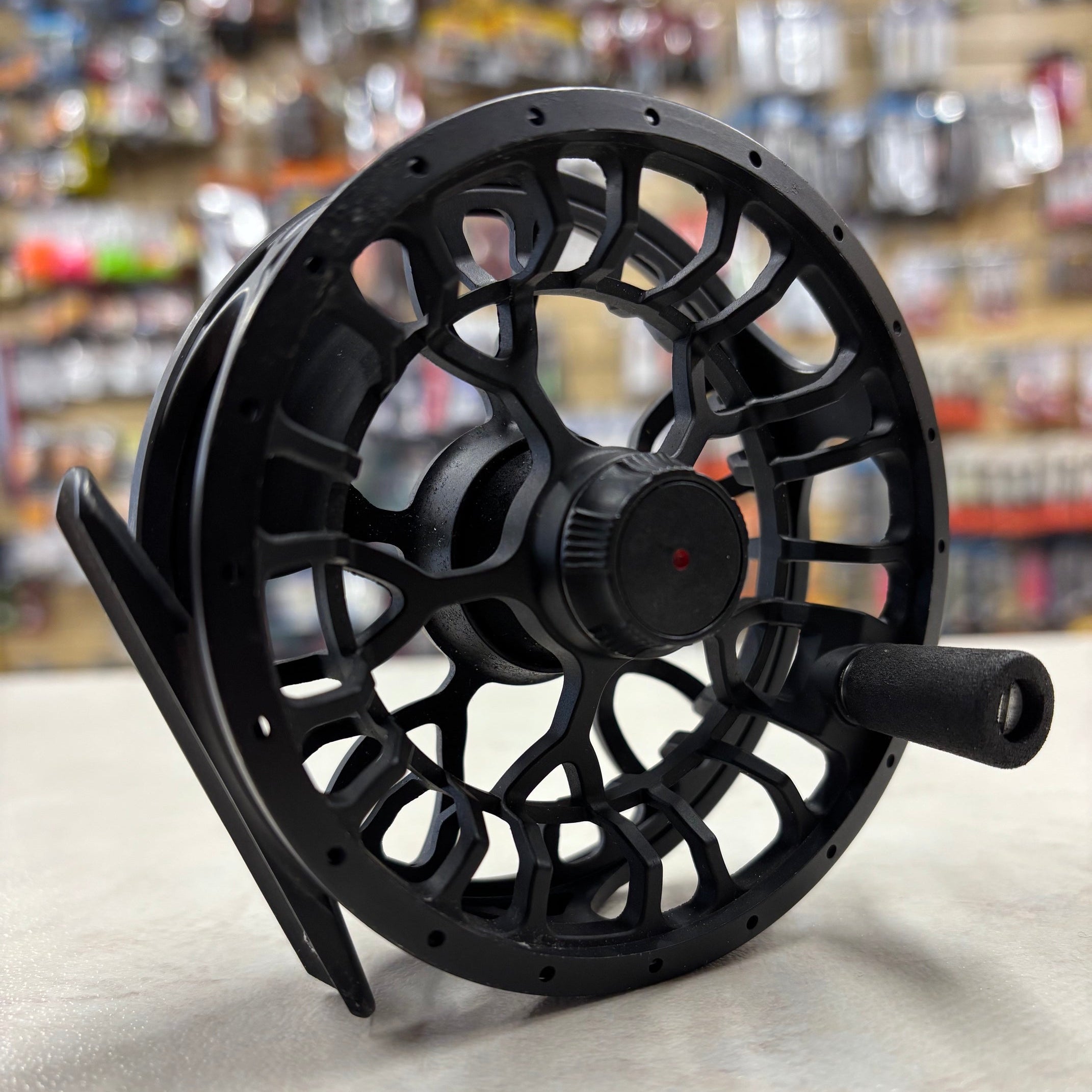 A black fly fishing reel with an open-frame design and a side handle, photographed on a light surface with a blurred store background.