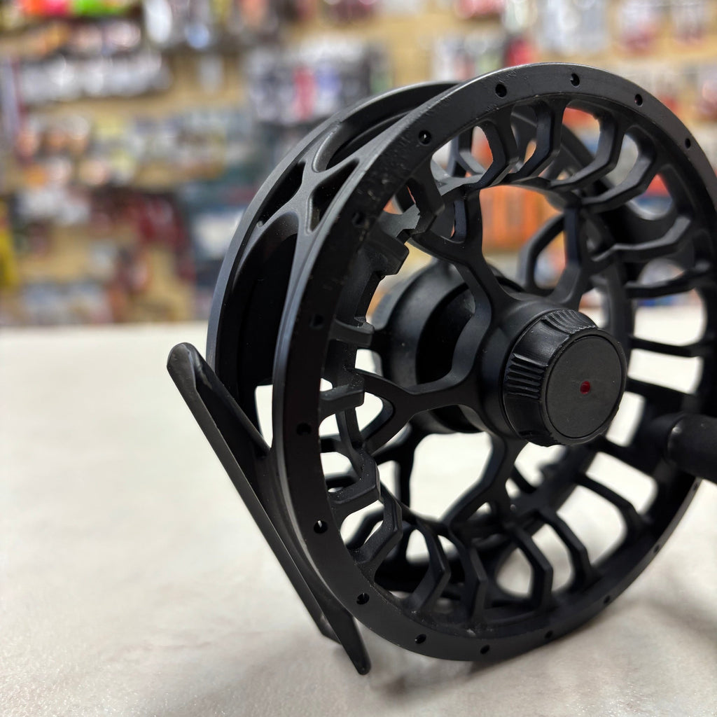A close-up of a black fly fishing reel with a textured spool, standing on a light surface with a blurred store background.